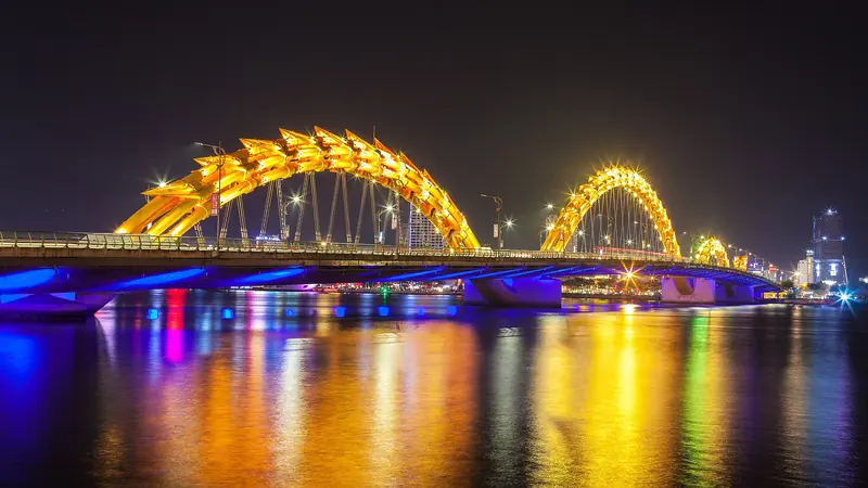 Dragon Bridge lit gold and blue at night, reflected in the Hàn River